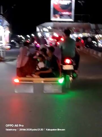 At night, a motorcycle pulls a pallet-like seat on a road. Multiple people are sitting on the seat, facing forward. Some of them are children. The seat has red and green lights. The motorcycle also has a red light. Other vehicles are on the road, including other motorcycles. There are buildings visible. A sign is also visible in the background.