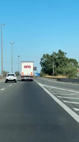 The video shows a perspective from inside a vehicle driving on a highway. Ahead, a white semi-trailer truck with "XPO" branding is visible in the right lane. To the left of the truck, on the shoulder, is a white car. On the right side of the highway, near the shoulder, there is a blue sign with stars and the word "België" written on it, indicating the border crossing into Belgium. The sky is clear and blue, and there are trees and vegetation along the side of the road. The highway has clear lane markings, and there are streetlights visible in the distance. The video is shot in a continuous forward motion, simulating a drive.