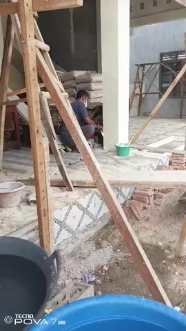 The video shows a construction site with scaffolding. In the first frame, a person wearing a blue shirt and a face mask is seen working on a wall. There are bags of materials and buckets around. The second to fifth frames show an outdoor scene with a person wearing a purple garment near a pile of dirt and construction materials. A road with buildings and some people on motorcycles is visible in the background. The overall environment appears to be in a town or city.