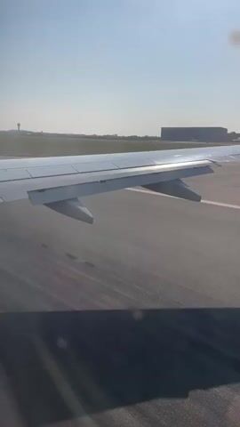 The video shows a view from an airplane window as it moves along a runway. The plane's wing is visible on the right side of the frame. The runway appears to be asphalt with white markings. In the background, there is a flat, open landscape with some buildings and a tall, slender structure that might be a control tower. The sky is clear and blue. The airplane is either taxiing or preparing for takeoff or landing.