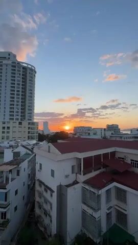 A vertical video shows a sunset over a city and the ocean. The sun is a bright, glowing orb near the horizon, casting a warm orange and yellow light across the sky. Wispy clouds are scattered across the sky, illuminated by the setting sun. In the foreground, there are several buildings, including a tall, modern high-rise on the left and smaller buildings with red roofs in the center and right. Trees are visible in front of some of the buildings. The ocean stretches out to the horizon, and distant islands can be seen. The overall scene is peaceful and serene.