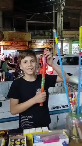 A young boy stands smiling on a street, holding a decorated tube. He then looks to the side, and the camera pans out. The scene is a busy street at night, with people walking by and cars parked along the road. There are shops lining the street, and goods, including shoes and sandals, are displayed for sale on a table. A woman wearing a hijab walks by.