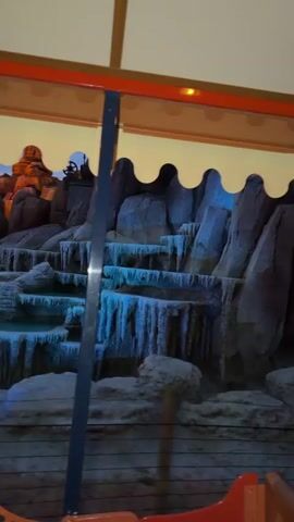 The video shows a view from inside a vehicle, possibly a train or boat ride, looking at a simulated rocky landscape with artificial waterfalls and vegetation. The scene is lit with dramatic lighting, including blue and red hues. A dark blue pole is visible in the foreground, along with the edge of the vehicle's interior, which appears to be red and orange. In the background, there are rocks and foliage, and the lighting suggests a twilight or nighttime setting. A person wearing a red, white, and blue plaid shirt is partially visible in the lower right corner, seated in the vehicle. The simulated environment appears to be part of an amusement park attraction, with the lighting and set design creating a theatrical effect. The video begins with a close-up of the rocks and then pans slightly, revealing more of the environment and the seated person. The overall impression is of an immersive, staged experience designed to evoke a sense of adventure or wonder.