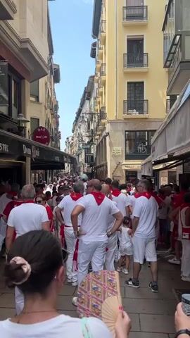 A long, narrow street is filled with a crowd of people. Most of the people are wearing white shirts and white pants, with red bandanas tied around their necks or waists. The buildings lining the street are tall and appear to be made of stone and plaster, with balconies and windows. The sky is visible between the buildings, and it appears to be a clear, sunny day. The people are walking in the same direction, creating a sense of movement and activity. Some people are looking at their phones, while others are talking to each other. The overall atmosphere seems to be festive and energetic.