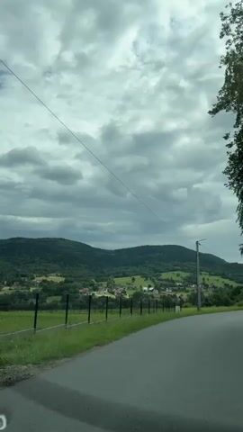 This is a vertical, first-person perspective video of a rural road winding through a valley. The sky is overcast with heavy gray clouds. On the left side of the road, there is a grassy verge with a black metal fence running along it. Beyond the fence, there are green fields and a scattering of houses, forming a small village nestled among rolling, tree-covered hills in the distance. A single utility pole with wires stretches diagonally across the frame. The road itself is paved and curves gently to the right. The overall impression is one of a natural, somewhat subdued landscape under a cloudy sky.