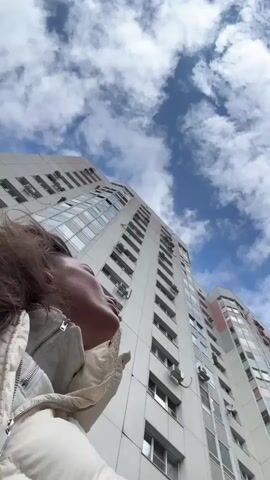A low-angle shot shows a woman with brown hair standing outside, looking up at a tall white building under a sky with scattered white clouds. The building has many windows and air conditioning units. The woman is wearing a light-colored jacket. Her hair is blowing slightly in the wind. The overall lighting is bright due to the daylight.