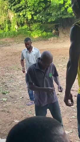 The video captures an outdoor gathering in a rural or semi-rural area, consistent with Ogbu, Nigeria, under bright daylight. The scene unfolds on a dirt ground bordered by lush green trees and foliage. In the foreground, a man is seated and actively playing a drum with sticks, with another drum visible beside him.

Several men are present, many of whom are moving rhythmically or dancing to the drum music. One man in a yellow sleeveless vest prominently displays "CELTIC 11" on his back, while another man wears a yellow sleeveless vest featuring a large printed image of a person's face. A man in a light grey polo shirt, blue jeans, and flip-flops, carrying a small grey cross-body bag, stands in the mid-ground, also moving. Another man in a dark grey polo shirt with a small green logo on the front is seen bending and moving his body. In the background, several children are visible playing in the open space. The atmosphere suggests a lively and communal cultural event or informal performance.
