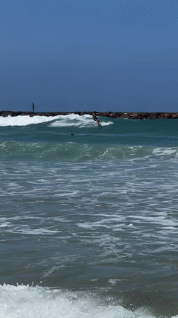 Surfer rides waves near Tel Aviv breakwater under clear skies