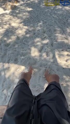 The video begins with a first-person perspective of someone sitting on a sandy beach, their black shorts and bare feet visible. The sand is white and textured, with dappled sunlight filtering through overhead trees, casting shadows. In the background, a few rocks and some indistinct playground equipment can be seen. The camera then pans up and slightly to the left, revealing more of the beach environment. Several beach umbrellas are set up, and a few people are visible in the distance, some sitting and one walking near the water. The ocean is a clear blue, and mountains form the backdrop on the horizon under a bright blue sky. The scene conveys a relaxed beach atmosphere.