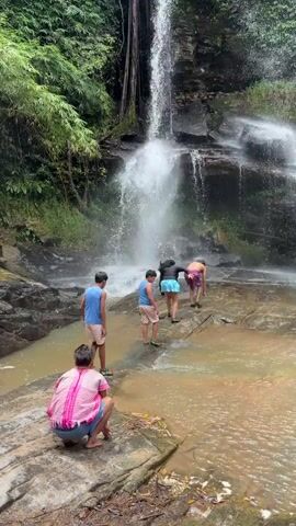 Five people are near a waterfall in a jungle. The waterfall is tall and cascades down rocky cliffs. The water is light brown. The people are wading through the water and climbing on the rocks. One person is in the foreground, crouching on a rock and facing away from the camera. Four other people are further back, walking towards the waterfall. The person closest to the waterfall is wearing a black top and blue shorts. The person next to them is wearing a pink and purple swimsuit. Two younger individuals in blue shirts and shorts are walking towards the waterfall. The environment is lush and green with trees and vegetation surrounding the waterfall.