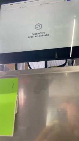 The video shows a close-up shot of a computer screen displaying "Scan of typ order en operatie" with a hexagon-like icon above it. Below the screen, there is a stainless steel surface with a green paper label partially visible on the left side. As the camera moves downwards, it focuses on the green paper label which is a product label with information printed in black, including the text "MBI De steenmeesters," "Geocorso," and dimensions of "80x40x4 cm," as well as "Rimini." In the reflection on the stainless steel, a person is visible holding a smartphone. The text on the label indicates that it relates to stone products.