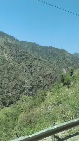 The video shows a view of a steep, forested mountainside with a river flowing at its base. A power line crosses the frame horizontally. The terrain is rugged and covered in dense green vegetation. The sky is clear and blue. The video appears to be filmed from a moving vehicle, likely a car or bus, due to the slight motion blur and the perspective. The time code indicates the video is very short, under 5 seconds.