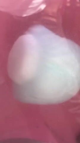 The video shows cotton candy being made in a pink cotton candy machine. The cotton candy is white with a hint of blue. A person holding a drink in a cup walks behind the machine. The camera focuses on the cotton candy as it is spun onto a paper cone. The background is out of focus.