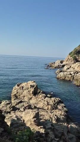The video shows a view of the ocean from a rocky coastline. The water is blue and calm, with gentle waves. The rocks are large and jagged, and covered in sparse vegetation. The sky is clear and blue, with no clouds. The sun is shining, casting shadows on the rocks. The scene is peaceful and serene.