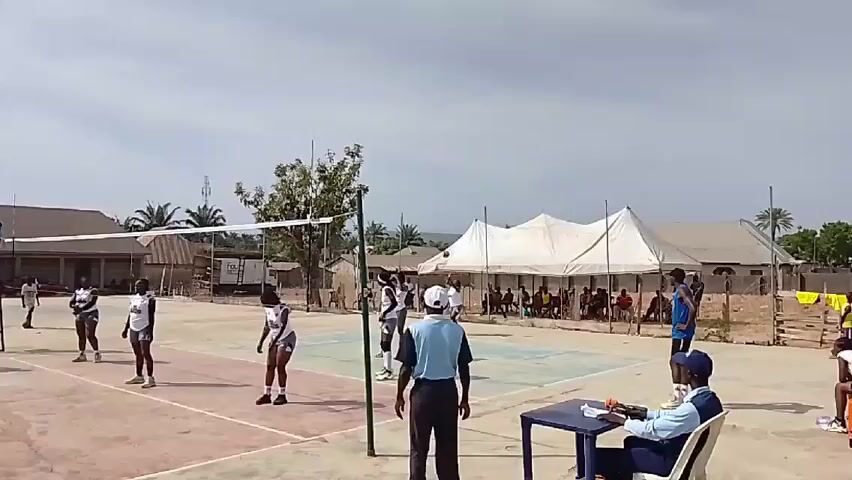 A volleyball game is in progress on an outdoor court. There are two teams of young women, one wearing blue and yellow uniforms and the other wearing white and black uniforms. The blue team is serving. The ball is in the air, with a blue player about to hit it. A referee stands on a raised platform near the net. Another referee sits at a table on the side, keeping score. Spectators are seated under a white tent in the background. The court is surrounded by buildings and trees. The sky is overcast.