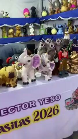 The camera pans across a vendor's booth displaying hand-painted plaster figures. There are two shelves. The top shelf is lined with a blue-and-white scalloped fabric. On the top shelf are a variety of animal figures, including owls, dogs, and pigs. There are also some cartoon characters, such as Stitch from the Disney movie "Lilo and Stitch" and a Baby Groot figure. The bottom shelf is lined with a white fabric. On the bottom shelf are a variety of animal figures, including elephants, owls, and eagles. There are also some anime characters, such as Goku from the "Dragon Ball" series. In the bottom right-hand corner of the video is a sign that says "Sector Yeso 2026."