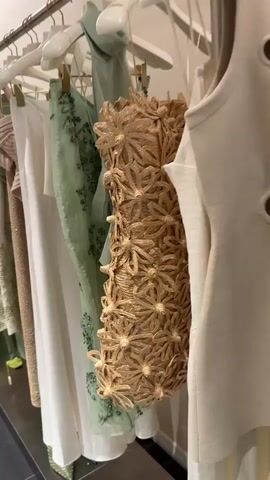 The video shows a rack of clothing with a person's hand interacting with one of the garments. There are multiple dresses hanging. The first dress that is fully visible is a pale green, semi-sheer fabric with floral embellishments. To the right is a textured beige dress. Further along the rack is a white dress. At one point a green, textured item can be seen tucked between the garments. The background is a bright white wall. The interaction consists of the person’s hand briefly moving the pale green dress.