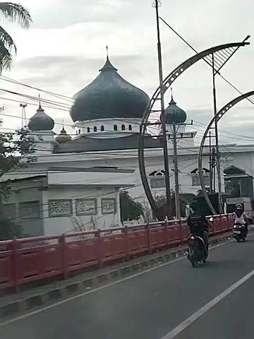 This video shows a street scene with a mosque in the background and several motorcycles driving on the road. The mosque has a large dark dome and several smaller domes. The building is white with ornate windows. There is a red railing along the side of the road. Two people on motorcycles are visible in the foreground, and another motorcycle is seen further down the road. The sky is overcast.