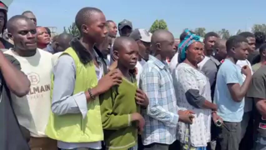 Here is a description of the video:

**Scene:** The video shows a medium-sized crowd of people, predominantly men, standing outdoors in what appears to be a public space, possibly a street or road. A few women are visible within the crowd.

**People:** The individuals appear to be of African descent. They are dressed in casual clothing, including T-shirts, jackets, and patterned shirts. Some individuals are looking at the camera, while others are focused elsewhere. One man is wearing a shirt with the word "BALMAIN" printed on it. A young boy, wearing a green sweater, is standing in the middle of the crowd.

**Environment:** The background suggests an urban or semi-urban setting with buildings, foliage, and what seems to be a road or open area. The weather appears to be sunny.

**Actions & Interactions:** The people are standing close together, suggesting a sense of unity or shared purpose. There seems to be some sort of gathering or demonstration occurring.

**Audio:** There is someone speaking in a language other than English.