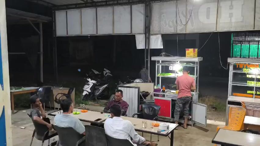 A casually lit nighttime scene depicts a modest open-air food stall or restaurant. Four men are seated around a table, casually dressed and engaged in conversation. On the opposite side, a man in a pink shirt appears to be preparing food at a mobile cooking station. Several motorcycles are parked near the back, adding to the informal atmosphere. The setting is simple, with basic furnishings and a focus on functional space.