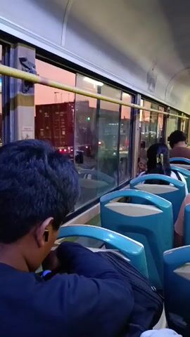 The interior of a city bus is shown from the perspective of a passenger seated towards the front. Other passengers occupy the seats in the row to the right, and the window beside the seats provides a view of the outside world. The scene captures a city street with cars and motorcycles driving, with buildings and a billboard visible against a backdrop of a pink and orange sky, indicating it might be dusk or dawn. The bus seats are predominantly turquoise. The person taking the video is wearing a dark blue shirt, and a black backpack rests in the empty seat next to them.