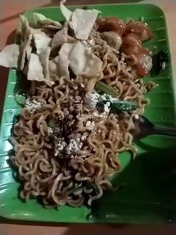 A fork is used to stir a plate of noodles with fried wonton, meat balls, and green vegetables on a green banana leaf-shaped plate.
