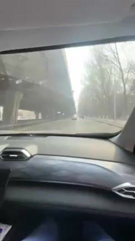 The video shows a car ride, filmed from the passenger seat. The view through the windshield reveals a city street with an overpass and trees along the side. Other cars are visible on the road. The camera briefly pans to the side window, showing more trees and a sidewalk. The interior of the car is visible, featuring a black dashboard and upholstery with silver accents. The passenger's legs and dark blue pants are partially visible in the bottom of the frame.