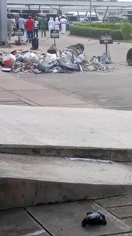 The video shows a pile of garbage in an outdoor area, possibly a parking lot. Several people are walking in the background, some wearing white uniforms. There are signs visible, one of which reads "NO PARKING". There are also parked vehicles in the background. The ground is paved.