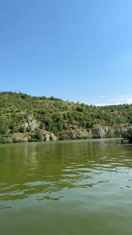 This media file captures a scenic view of a lake with lush green hills in the background under a clear blue sky. The water is a vibrant green, reflecting the sunlight and creating ripples on its surface. The hills are densely covered with trees and interspersed with rocky outcrops, suggesting a natural, possibly untamed, landscape. In the distance, a few power lines can be seen against the sky, hinting at human presence in the area.

The activity taking place is a leisurely boat ride on the lake, as evidenced by the perspective of the camera seemingly moving through the water. The presence of a hand making a peace sign in the foreground suggests a moment of enjoyment and captured memory. The time of day appears to be midday or early afternoon, given the bright sunlight and clear sky. The weather seems pleasant and warm, ideal for outdoor activities.

While no text is explicitly visible, the location context indicates that this scene is in Lukovit, Bulgaria. The overall mood conveyed is one of tranquility and natural beauty, with a touch of playful interaction from the person filming. The combination of the calm water, verdant landscape, and clear sky creates a peaceful and inviting atmosphere.