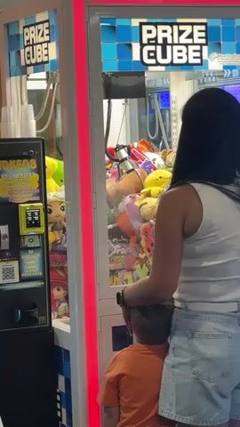 A woman and a young boy stand in front of a prize cube claw machine. The machine is illuminated with blue, purple, green, and red LED lights along its edges. The front of the machine displays the words "PRIZE CUBE" in large letters. To the left of the claw machine is a token dispenser with a digital screen displaying "TOKEN PRICES" and various token options. The claw machine's interior is filled with an assortment of plush toys, including cartoon characters and animals. The woman, with long dark hair, is wearing a white tank top and denim shorts, and the boy, wearing an orange t-shirt, is standing close to her, looking into the machine. The lighting suggests an indoor arcade or entertainment venue.