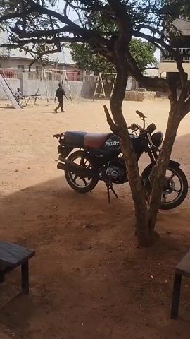 The video shows a schoolyard with a black motorcycle parked under a tree. The motorcycle has the word "PILOT" written on its side. A person is walking in the background across the sandy schoolyard. There are benches and playground equipment visible. The school building is in the background. The video is taken from a slightly low angle, panning to show different parts of the yard.