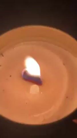 A close-up, slightly shaky shot shows a lit candle in a round, metallic container. The wax is a light beige color and has some small dark particles on its surface. The flame is bright with a blue and reddish base transitioning into white and yellow at the top. The camera angle is directly above, looking down into the candle. The background is dark and out of focus.