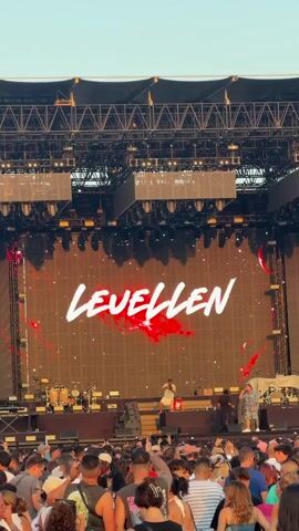 A wide shot captures a lively outdoor music festival. A large LED screen displays the word "LEVELLEN" in bold white lettering with red accents. On stage, two performers are visible. The foreground is filled with a dense crowd of people, mostly facing the stage. The lighting suggests it is daytime or early evening. The overall atmosphere appears energetic and engaged.
