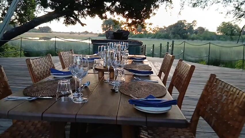 A wooden table, set for a meal, stands on a wooden deck. Each place setting has a charger plate made of woven material, a white plate topped with a blue napkin, a wine glass, a smaller glass, a spoon, and a knife. Woven-backed wooden chairs surround the table. In the background is a body of water, with green reeds and grass separating it from the deck. Beyond the water are silhouetted trees under a sunset sky.  A small, green tiled structure sits off to the right. A large tree with branches reaching over the deck partially obscures the view.