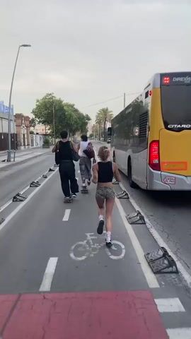 The video shows a person running on a street while three other people on skateboards follow behind. A yellow bus is driving alongside them. The street has a bike lane marked with a bicycle symbol and white lines. The bus has the word "CITARO" written on its back. There are some buildings and trees in the background. The sky is overcast.