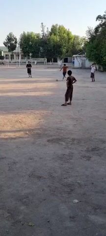 The video shows a group of children playing soccer in an outdoor schoolyard in Havaskor, Tajikistan. The ground is a dirt field, and there are trees and school buildings in the background. The sun is shining, suggesting it's daytime, likely afternoon given the long shadows. The children appear to be having fun, engaged in a casual game of soccer. One child in a brown shirt and pants is dribbling the ball, while others are positioned around the field, ready to receive a pass or defend. A soccer goal is visible in the distance. The overall atmosphere is one of youthful energy and activity in a rural or semi-rural setting. There is no visible text in the video.