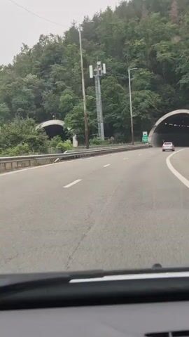 This video shows a car driving on a highway towards a large tunnel entrance. The highway is paved with clear lane markings and is bordered by a metal guardrail. On the left side of the highway, there is a tall cell tower with antennas and a lamppost. Dense, green trees cover a steep hillside to the left and behind the tunnel. Another tunnel entrance is visible further in the distance on the left. A white car is ahead of the viewer's car, driving towards and entering the tunnel on the right. A green sign with white text and an icon of a tunnel is visible on the left side of the road before the tunnel entrance. The text on the sign appears to read "TUNNEL" and "795 m". The sky is overcast, suggesting a cloudy day. The overall scene is one of travel through a scenic, wooded area.