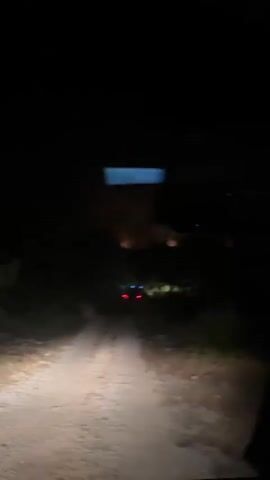 The video shows a first-person perspective of driving down a dirt road at night. The headlights of the vehicle illuminate the rough, uneven path ahead, bordered by dark trees and brush. In the distance, through the trees, a large orange glow is visible, suggesting a fire. There are no people clearly visible, but there are some red and blue lights in the distance near the glow, possibly indicating emergency vehicles or other activity related to the fire. The sky is dark and the overall atmosphere is one of urgency or concern due to the fire in the distance.