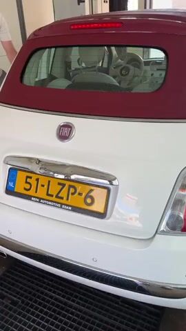The video shows a white Fiat 500 convertible at an automated car wash. The camera pans across the back and side of the car, highlighting its license plate, taillights, and the retractable roof. We then see the car being guided through the car wash by a track, with large brushes and cleaning equipment visible. The car wash appears to be outdoors, with a paved surface and some greenery in the background. The video also shows the car's interior, including the steering wheel and seats. The overall impression is that of a car undergoing a routine wash.