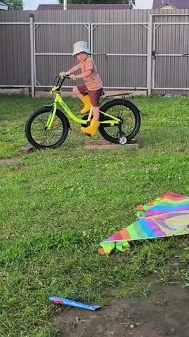 A young child wearing a light-colored bucket hat, a patterned t-shirt, brown shorts, and yellow rain boots rides a bright yellow bicycle with training wheels. The bicycle is positioned on two rectangular wooden blocks placed on a grassy lawn. The child is gripping the handlebars and appears to be pedaling. In the foreground, a colorful kite lies on the grass, and a blue and red object is partially submerged in water. A metal fence with vertical panels stands in the background, behind which houses are visible.