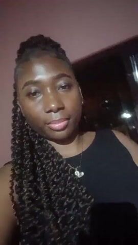 A young woman with dark skin and braided hair styles poses for a selfie-style video. She is wearing a black tank top and a necklace. The background appears to be a nightclub or bar, with a red and black sign visible in some frames. The sign says "REDBOX THE GOOD LIFE". The lighting is dim, and the woman appears to be posing for the camera, subtly moving her head and arm. There is music playing in the background.