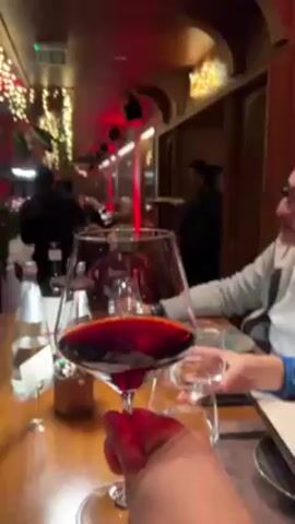 The video shows the interior of a restaurant, likely during the evening or nighttime. The scene opens with a view focused on a glass of red wine on a wooden table. Other glasses, water bottles, and place settings are also visible.  The camera then pans around, showing other diners seated at the table, a waiter moving in the background, and interior design elements such as plants and decorative lights. A large portrait or image on the wall is reflected in the window. The camera moves to show a long dining table set for a meal, located near a window that overlooks a city at night.