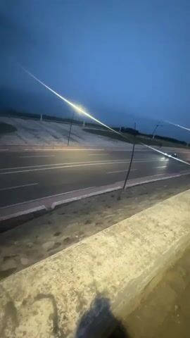 The video captures a dark-colored sedan driving on a multi-lane road at dusk or night in Fnideq, Morocco. The car, with its headlights illuminated, moves from left to right across the frame. Streetlights are visible, casting bright, elongated streaks of light across the scene, indicating the low-light conditions. The sky is a deep blue, suggesting either twilight or very early night. In the foreground, a concrete barrier or wall is visible, implying the footage was taken from an elevated viewpoint overlooking the road. The road has clear lane markings, and there are additional structures that appear to be part of the roadside or infrastructure in the background. No people are visible, and there is no discernible text within the video. The atmosphere is quiet and serene.