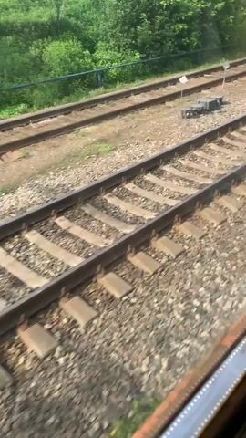 The video shows a view from a train window looking out at adjacent train tracks. The tracks are made of metal rails and gravel ballast. Green foliage and trees are visible on the left side of the frame, with a fence line separating them from the tracks. There are multiple sets of parallel train tracks. In the distance, there are some buildings, a fence, and what appear to be train signals or markers. The camera is moving, indicating the train is in motion. The lighting suggests it is daytime. There are no people or animals visible in the frame.