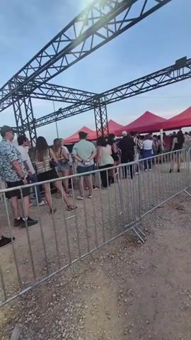 The video shows a line of people waiting under a metal structure. Red tents are visible in the background. People are dressed in casual clothing, and some are holding backpacks. The ground is dirt and gravel. The sky is overcast.