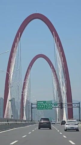 The video shows a wide, multi-lane highway with cars driving on it. The highway is elevated, suggesting it's a bridge or overpass. In the background, two massive, arched structures, colored a deep red and supported by numerous white cables, dominate the skyline. These arches appear to be part of a suspension bridge. Green highway signs with white lettering are visible above the road, indicating directions and distances to various locations. The sky is clear and blue, suggesting a sunny day. The overall scene conveys a sense of modern infrastructure and transportation.