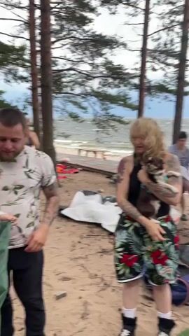 A group of people are camping on a sandy beach next to a body of water, possibly a lake or ocean. The sky is overcast. Several tents are set up, and camping gear is scattered around. In the foreground, a person with blonde hair and tattoos is holding a small dog. To their left, another person with tattoos is partially visible. To their right, a man in a green shirt is interacting with a green tent. Further back, a woman is standing near a tent, and another couple is seen near a kayak or canoe. The overall scene depicts a casual camping environment with various activities taking place.