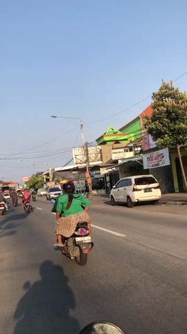 A first-person perspective video shows a person riding a motorcycle on a road. The rider is wearing a green shirt and a plaid skirt. In front of the rider, other motorcycles and a white van are visible on the road. The scene appears to be in a populated area with buildings and trees lining the street. The sky is clear and blue, suggesting daytime. The sun is low in the sky, casting long shadows.