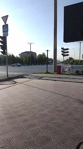 The video pans right across a pedestrian walkway paved with decorative concrete tiles. To the left, a traffic light with a pedestrian crossing signal is visible. A road intersection is in the background with a few cars and trees. In the far distance, buildings and a tall utility pole can be seen. The sky is clear and bright, suggesting it is daytime.