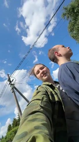 A female and a male are captured in a low-angle, moving shot from a selfie perspective. The female, positioned on the left and closer to the camera, wears a green camouflage jacket and has light brown hair with strands blowing in the wind. She looks upwards and to her right. The male, positioned behind and to the right of the female, wears a dark blue long-sleeved shirt and light grey pants. He has short brown hair and looks directly upwards. The background displays a bright blue sky with scattered white clouds, power lines, utility poles, green trees, and distant buildings, suggesting an outdoor, sunny environment. The camera slightly wobbles, indicating movement.